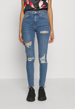 Slim-fit, blå denimjeans med slitna detaljer, som har flera revlar på knäna. Tillsammans med svarta sneakers och en blommig topp.
