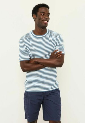 Lächelnder Mann mit kurzen lockigen Haaren, der ein blau-weiß gestreiftes kurzärmeliges Hemd und dunkelblaue Cargo-Shorts trägt, die Arme verschränkt, blickt nach rechts.