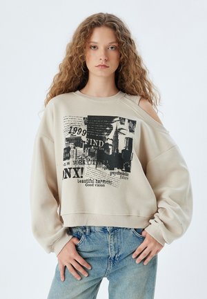 Sudadera beige con hombros descubiertos, que presenta estampados gráficos en negro, incluyendo texto y un horizonte de ciudad, corte relajado y dobladillo acanalado.