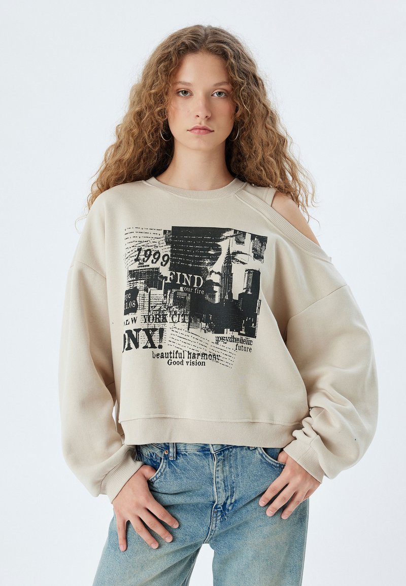 Felpa beige con spalle a strappo, caratterizzata da stampe grafiche nere che includono testo e uno skyline, vestibilità comoda e orlo a coste.