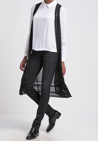Černý dlouhý vestový kabát s krajkovými detaily, který je nošený přes bílou košili s límcem a černé slim-fit kalhoty, doplněný o lesklé černé boty.