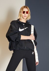 Črna vetrovka s visokim ovratnikom in velikim belim logotipom Nike, v kombinaciji s črnimi leginami in belo naramnico s studenci. Rdeče sončne očala.