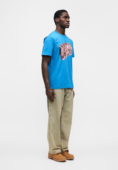 Μπλε βαμβακερό μπλουζάκι με γραφικό σχέδιο, συνδυασμένο με μπεζ παντελόνι με κάθετες ρίγες και καφέ μπότες, που παρουσιάζεται σε όρθιο σχήμα.