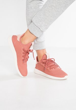 Person iført lysegrå joggingbukser og koralrøde mesh-sneakers med hvide såler, stående på et hvidt gulv.