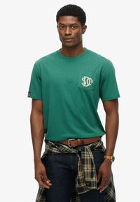 Camiseta verde de manga corta hecha de una tela suave, con un bolsillo en el pecho que presenta un diseño gráfico. Combinada con unos jeans oscuros y una camisa de cuadros atada a la cintura.