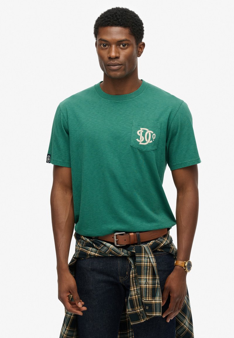 Camiseta verde de manga corta hecha de una tela suave, con un bolsillo en el pecho que presenta un diseño gráfico. Combinada con unos jeans oscuros y una camisa de cuadros atada a la cintura.