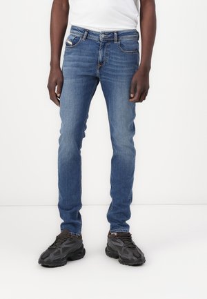 Homme portant un jean slim bleu et des chaussures de sport noires, debout devant un fond blanc uni.
