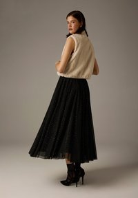 Cremefarbene Kunstpelzweste mit hohem Kragen, kombiniert mit einem langen schwarzen Tüllrock mit verstreuten silbernen Punkten und schwarzen Riemchen-High Heels.