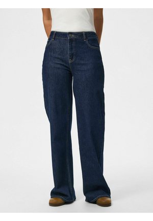 Persoon die donkerblauwe wijde jeans, een wit topje en bruine schoenen draagt, staand tegen een effen lichte achtergrond.