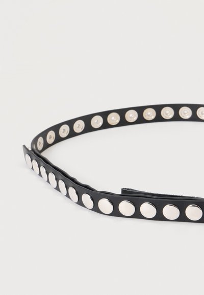Ceinture en cuir noir avec une rangée de clous ronds argentés régulièrement espacés, présentée enroulée sur un fond clair et uni.
