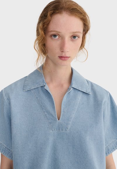 Jeune femme aux cheveux roux clair et aux yeux bleus portant une chemise en denim bleu clair avec un col en V profond sur un fond uni.