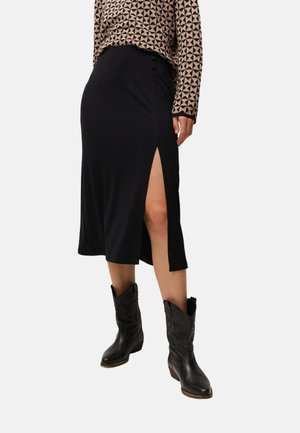 Mujer con una falda midi negra con una abertura alta en el muslo, top de manga larga con estampado y botas vaqueras negras hasta media pantorrilla, de pie contra un fondo blanco.