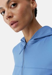 Ljusblå zip-up huvtröja i slät tyg, med dragsko i huvan och en strömlinjeformad design. Närbild på halsringningen och ansiktet.