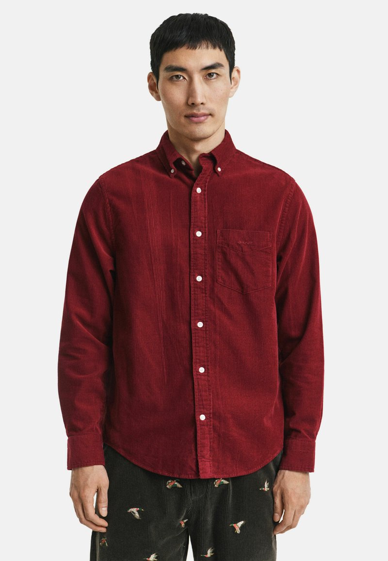 Chemise boutonnée en velours côtelé rouge avec une poche poitrine, dotée de boutons blancs et d'une texture subtile. Manches longues et ourlet arrondi.