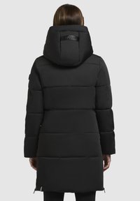 Eine Frau von hinten, die einen langen schwarzen gepolsterten Wintermantel mit einer großen Kapuze und horizontalen Steppnähten trägt.