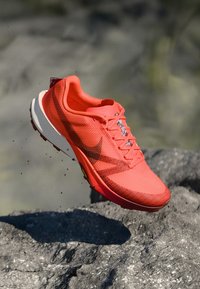 Rød atletisk sko med åndbart mesh-overdel, reflekterende accenter og en tekstureret gummisål. Designet inkluderer et fremtrædende swoosh-logo.