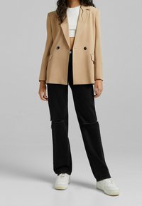 Beżowy dwurzędowy żakiet z wyłogami, dwa przednie kieszenie, w połączeniu z czarnymi podartymi dżinsami i białymi trampkami. Płaskie tło.