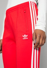 Crvene Adidas hlače imaju bijele pruge sa strane, džep sa zatvaračem i mali bijeli logo iznad koljena. Glatka, atletska tekstura tkanine.
