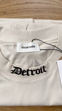 Gaiša béžā t-krekls ar apaļu kakla izgriezumu, mīkstu audumu un melnu izšūtu "Detroit" uz priekšpuses. Zīmola etiķete redzama iekšpusē.
