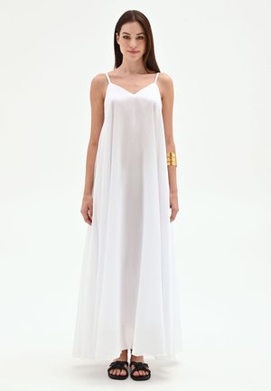 Femme aux longs cheveux bruns portant une robe maxi blanche sans manches et des sandales noires, debout devant un fond blanc uni.