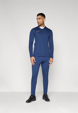 Mornarska atletska trenirka z zip-up jakno, dolgimi rokavi in ujemajočimi hlačami. Ima bele detajle in Nike logotip na prsih ter stegnu.