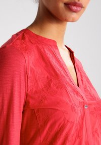 Blouse rouge avec un décolleté en V et des broderies florales. Présente des manches longues et un bouton décoratif avec un accent de drapeau. Texture lisse.