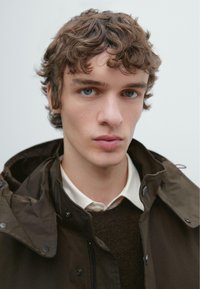 Brązowa, wodoodporna kurtka z szerokim kołnierzem i napami, noszona przez młodego mężczyznę na ciemnym swetrze i białej koszuli z kołnierzykiem.