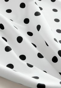 Tissu blanc à pois noirs irréguliers, délicatement plié pour montrer la texture et le motif.