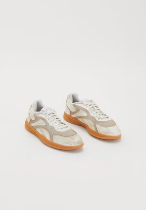 Beige sneakers with a textured, mixed-material design. Features white laces, a rubber orange sole, and geometric accents on the upper.