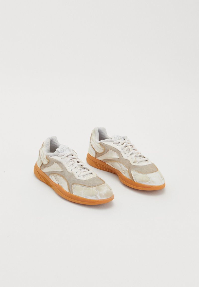 Beige sneakers med en teksturert, blandet materialedesign. Har hvite snøringer, en gummioverflate i oransje og geometriske detaljer på overdelen.