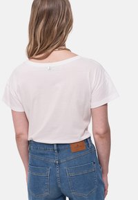 Camiseta de algodón blanca con mangas cortas y un corte holgado. Presenta un pequeño detalle de botón en la parte posterior, combinada con jeans de mezclilla azules.