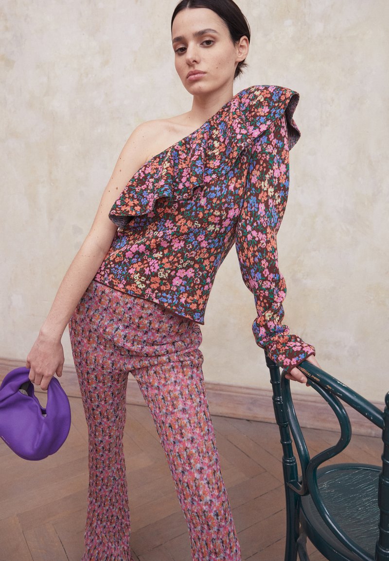 Top com padrão floral e franzidos em um ombro, combinado com calças texturizadas em tons de rosa e roxo; segurando uma bolsa roxa.