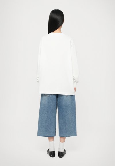 White long-sleeve T-shirt with a relaxed fit, paired with cropped blue denim pants featuring a frayed hem and back pockets. Black footwear completes the look.