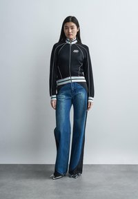 Jauna moteris stovinti vilkinti juodą striukę su užtrauktuku, dryžuotomis rankogaliais ir dvispalviais plačiomis kelnėmis bei sidabrinės spalvos aštriais bateliais.