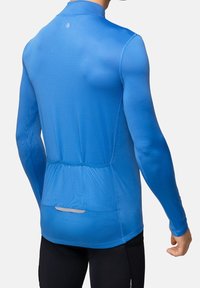 Camisa de deporte de manga larga en azul brillante, con una textura suave, diseño ajustado y un bolsillo trasero con detalles reflectantes.