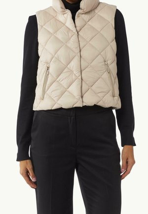 Veste matelassée beige sans manches avec poches zippées portée par-dessus un haut noir à manches longues et un pantalon noir.