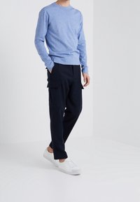 Jasnoniebieski sweter z długim rękawem, granatowe spodnie cargo z bocznymi kieszeniami oraz białe buty sportowe. Codzienny strój w minimalistycznym otoczeniu.