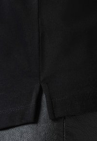 Camiseta negra con textura acanalada, con una abertura lateral y costuras limpias. La tela parece suave sobre un fondo de cuero.