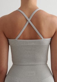 Top deportivo gris con relieve que cuenta con tirantes en cruz y un diseño ajustado, con un logo grabado en la banda. El material parece elástico.