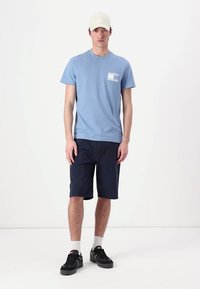 Lys blå bomull T-skjorte med brystlomme, marineblå shorts, svarte sko og en beige caps; står på en ensfarget bakgrunn.