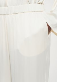 Tejido color crema con una textura suave, que presenta una cinturilla elástica y bolsillos laterales. Diseño minimalista con un brillo sutil.