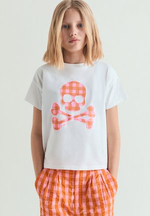 Giovane con capelli biondi fino alle spalle che indossa una t-shirt bianca con teschio e ossa incrociate a quadretti arancione-rosa e pantaloncini abbinati a quadretti.
