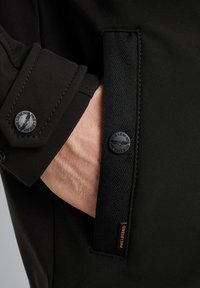 Zwarte jas met een close-up van een zak en knoopdetail. Bevat textuurstof, een geweven accent en een logo-label.