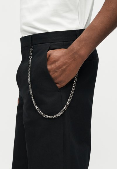 Χέρι που ακουμπά στην τσέπη μαύρου παντελονιού με προσαρτημένη ασημένια αλυσίδα, που φοράει λευκό πουκάμισο, με φόντο απλό λευκό.