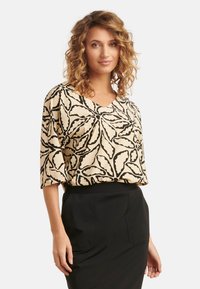 Beige blouse met zwart loofpatroon, korte mouwen en een v-hals ontwerp. Draag met een zwarte rok, wat de relaxte pasvorm van de top benadrukt.