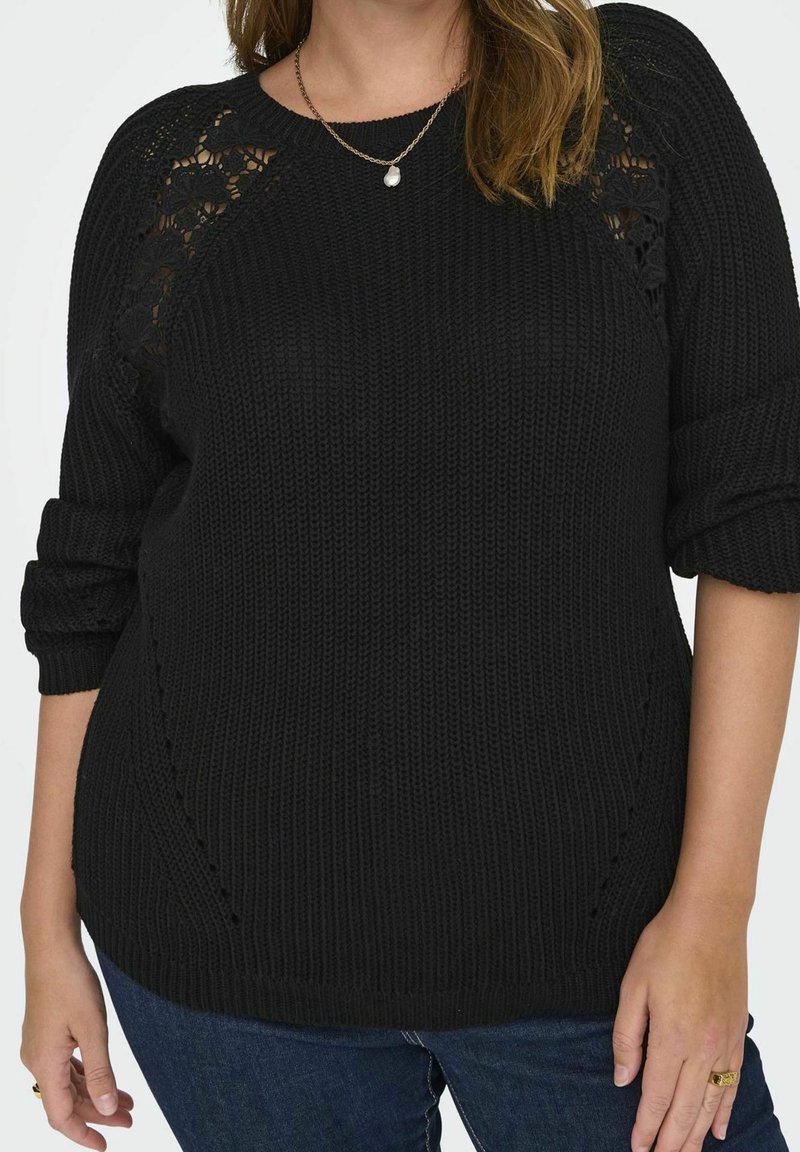 Svart stickad tröja med en strukturerad mönster och öppna spetsdetaljer på axlarna. Har uppvikta ärmar och en avslappnad passform.