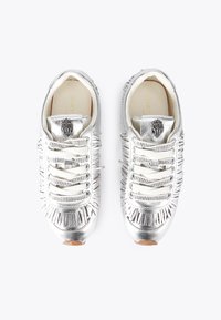 Coppia di scarpe da ginnastica argentate metalliche con lati frangiati, lacci bianchi e emblema nero sulla linguetta, viste dall'alto su sfondo bianco.