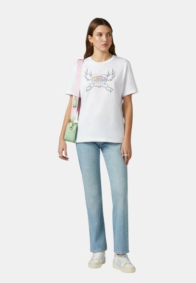 T-shirt bianco in cotone con grafica multicolore raffigurante ali e un cuore, abbinato a jeans a gamba dritta blu chiaro e sneakers con fantasia.