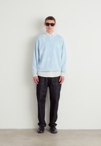 Sudadera azul claro con patrones de nubes, llevada sobre una camisa blanca con cuello, combinada con pantalones de mezclilla oscuros y zapatos negros, modelo usando gafas de sol.