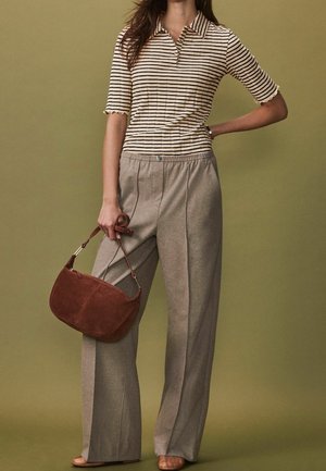 Donna con top a maniche corte a righe beige e pantaloni ampi taupe, che tiene una borsa in camoscio marrone, in piedi davanti a uno sfondo oliva.